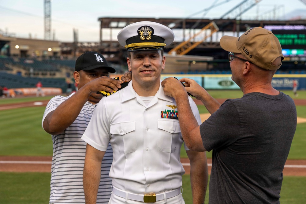 A U.S. Navy officer in white dress uniform is having insignia or rank devices pinned onto his shoulders by two civilians at what appears to be a baseball stadium. This appears to be a military promotion or commissioning ceremony conducted on a baseball field. The officer is smiling while two men simultaneously pin items to each of his shoulders.