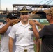 A U.S. Navy officer in white dress uniform is having insignia or rank devices pinned onto his shoulders by two civilians at what appears to be a baseball stadium. This appears to be a military promotion or commissioning ceremony conducted on a baseball field. The officer is smiling while two men simultaneously pin items to each of his shoulders.
