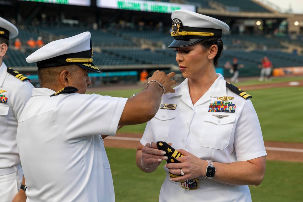 A military promotion ceremony is taking place on a baseball field, where a senior U.S. Navy officer is pinning new rank insignia on a female officer. The female officer is holding what appears to be shoulder board epaulettes with gold star insignia, suggesting a promotion to a flag or senior officer rank. The ceremony is occurring at what appears to be a professional baseball stadium during a game or event.