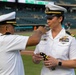A military promotion ceremony is taking place on a baseball field, where a senior U.S. Navy officer is pinning new rank insignia on a female officer. The female officer is holding what appears to be shoulder board epaulettes with gold star insignia, suggesting a promotion to a flag or senior officer rank. The ceremony is occurring at what appears to be a professional baseball stadium during a game or event.
