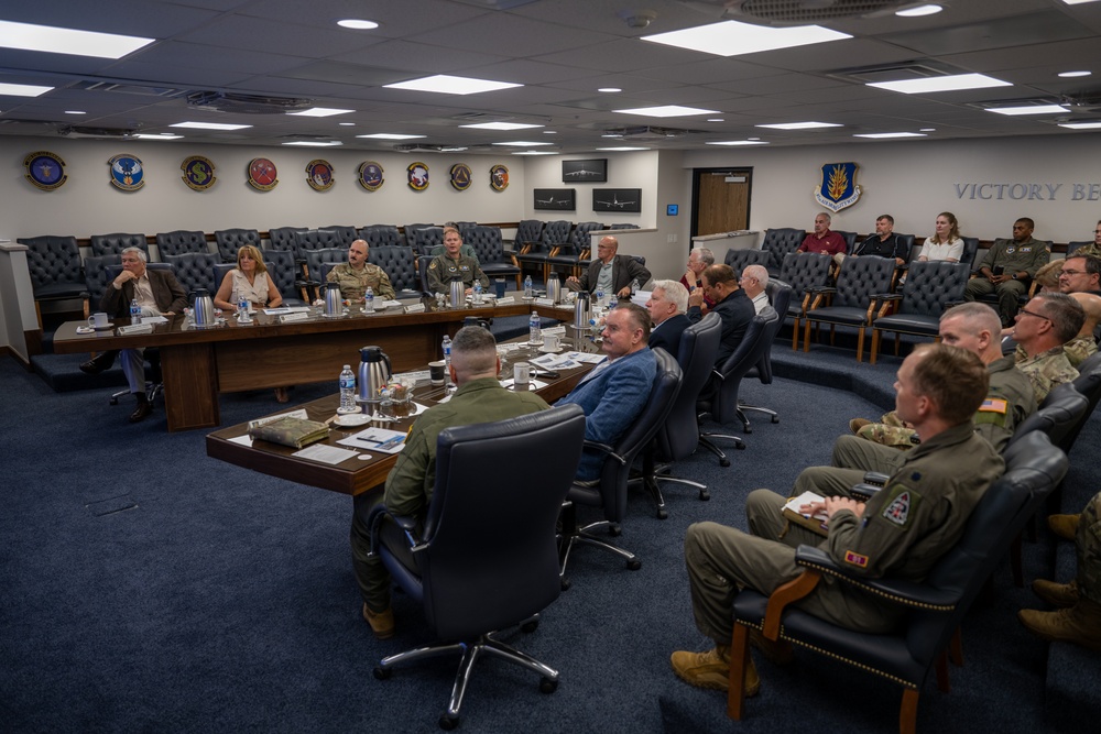 A formal briefing or meeting is taking place in a U.S. Air Force conference room, with a mix of military personnel in flight suits and Army uniforms alongside civilian attendees. The room displays multiple squadron patches and insignia on the walls, along with the text 'VICTORY BE...' (partially visible) and a U.S. Air Force emblem. Participants are seated around a large curved conference table and in chairs along the perimeter of the room.