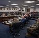 A formal briefing or meeting is taking place in a U.S. Air Force conference room, with a mix of military personnel in flight suits and Army uniforms alongside civilian attendees. The room displays multiple squadron patches and insignia on the walls, along with the text 'VICTORY BE...' (partially visible) and a U.S. Air Force emblem. Participants are seated around a large curved conference table and in chairs along the perimeter of the room.