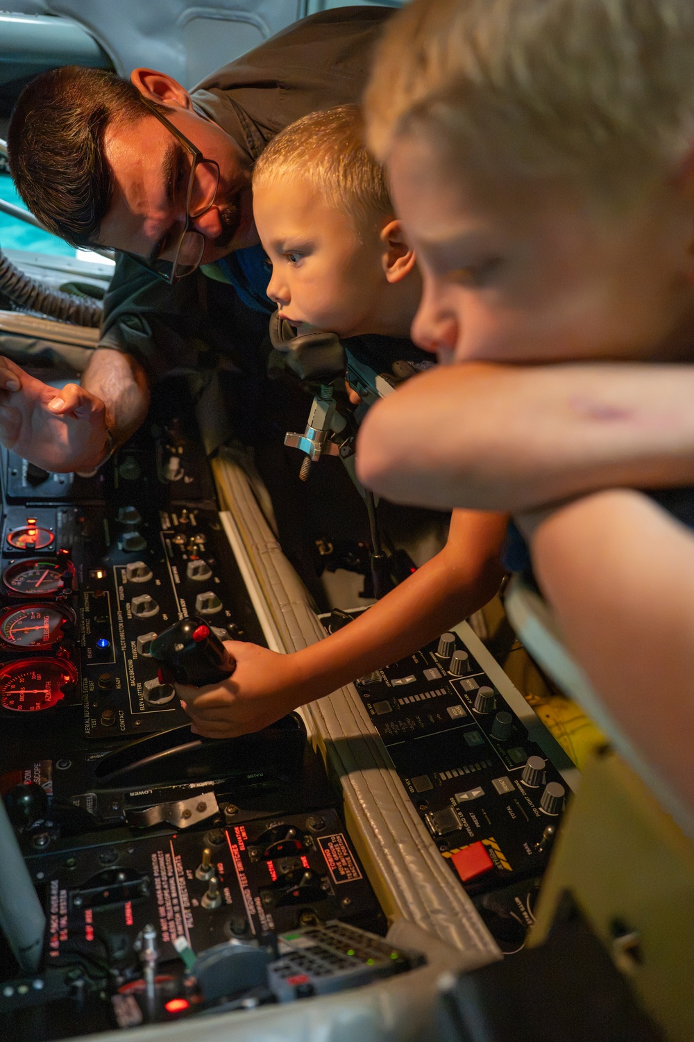 An adult male wearing glasses is showing two young blonde children the interior controls of what appears to be a military aircraft cockpit or crew station. One child is seated in a crew seat and is gripping a joystick/control handle while intently examining the instrument panel. The scene appears to be a guided tour or open-house event allowing public access to the aircraft.