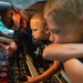 An adult male wearing glasses is showing two young blonde children the interior controls of what appears to be a military aircraft cockpit or crew station. One child is seated in a crew seat and is gripping a joystick/control handle while intently examining the instrument panel. The scene appears to be a guided tour or open-house event allowing public access to the aircraft.