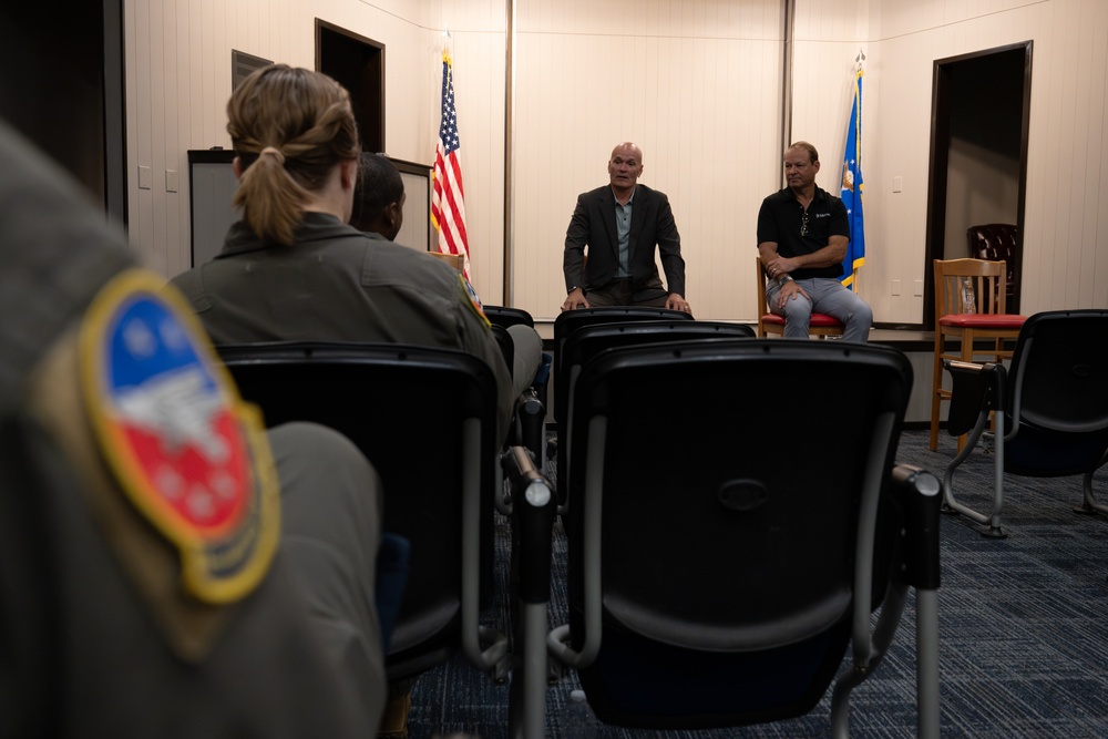 A small briefing or meeting is taking place in what appears to be a military facility room. Two civilian men are seated at the front facing an audience of military personnel in flight suits. An American flag and what appears to be a US Air Force flag are visible in the background.