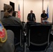 A small briefing or meeting is taking place in what appears to be a military facility room. Two civilian men are seated at the front facing an audience of military personnel in flight suits. An American flag and what appears to be a US Air Force flag are visible in the background.