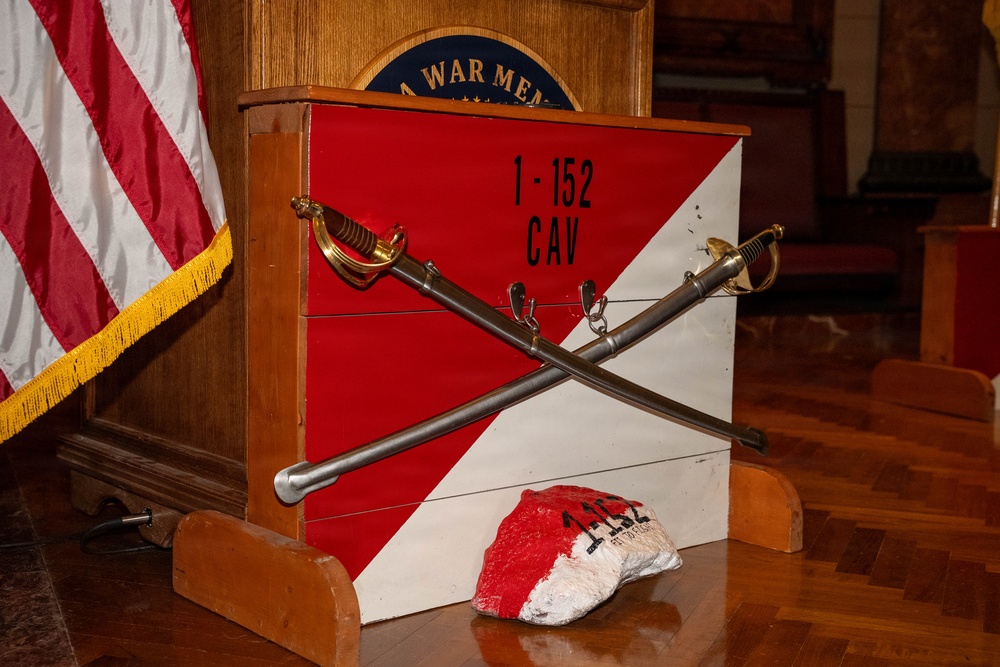A military unit display for the 1-152 CAV (1st Squadron, 152nd Cavalry Regiment) is set up in what appears to be a war memorial hall. Two crossed cavalry sabers are mounted on a red-and-white diagonal unit guidon box, with a painted rock bearing the unit designation placed in front. An American flag with gold fringe is visible to the left, and a podium with a partially visible 'WAR MEM[ORIAL]' seal is in the background.