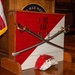 A military unit display for the 1-152 CAV (1st Squadron, 152nd Cavalry Regiment) is set up in what appears to be a war memorial hall. Two crossed cavalry sabers are mounted on a red-and-white diagonal unit guidon box, with a painted rock bearing the unit designation placed in front. An American flag with gold fringe is visible to the left, and a podium with a partially visible 'WAR MEM[ORIAL]' seal is in the background.