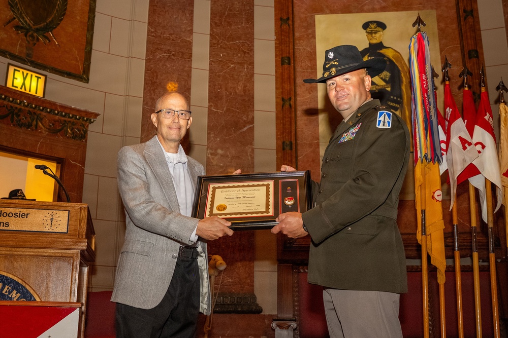 Two men are posing together while jointly holding a framed Certificate of Appreciation during what appears to be a formal military ceremony. One man is in civilian attire (gray blazer) and the other is in U.S. Army dress uniform with a campaign hat, medals, and unit patches. The setting appears to be a formal hall with military flags, a portrait of a military officer, and a podium visible in the background.