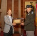 Two men are posing together while jointly holding a framed Certificate of Appreciation during what appears to be a formal military ceremony. One man is in civilian attire (gray blazer) and the other is in U.S. Army dress uniform with a campaign hat, medals, and unit patches. The setting appears to be a formal hall with military flags, a portrait of a military officer, and a podium visible in the background.
