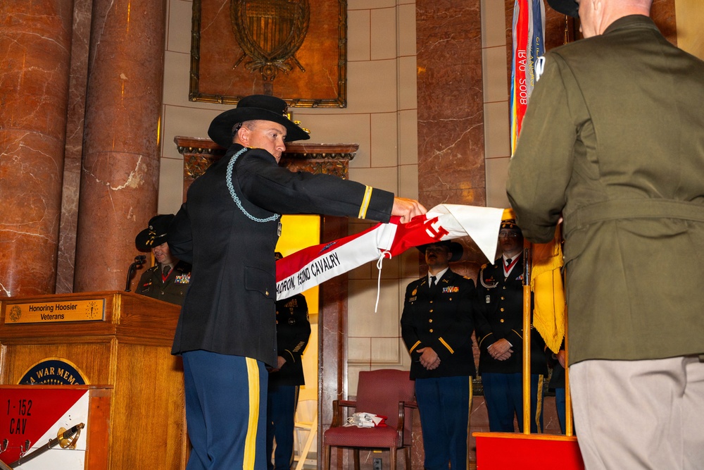 A military ceremony is taking place inside what appears to be the Indiana War Memorial, as indicated by the podium signage reading 'Honoring Hoosier Veterans' and 'War Memorial.' A soldier in dress uniform and cavalry Stetson hat is unfurling or presenting a unit guidon banner reading 'Squadron, 152nd Cavalry.' Several uniformed soldiers stand at attention in the background as part of a formal color guard or activation/inactivation ceremony.