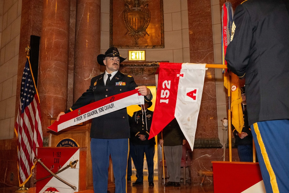 A U.S. Army soldier in dress uniform and cavalry black Stetson hat is holding an organizational streamer or guidon banner reading 'A Troop, 1st Squadron, 152nd Cavalry' during a formal military ceremony. The event appears to be taking place inside a war memorial building, with marble columns, an American flag, and military unit colors visible. Additional soldiers and civilians are present in the background.