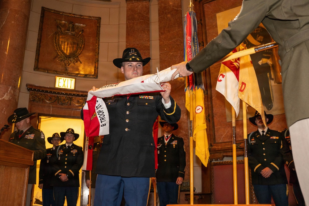 A formal U.S. Army military ceremony is taking place in an ornate indoor venue, likely a government or historic building. A soldier wearing a cavalry black Stetson hat and dress uniform is receiving or presenting a unit guidon/flag, with the text 'B TROOP' and 'CAVALRY' visible on the furled flag. Multiple soldiers in dress uniforms and cavalry hats are standing at attention in the background near additional unit colors and guidons.