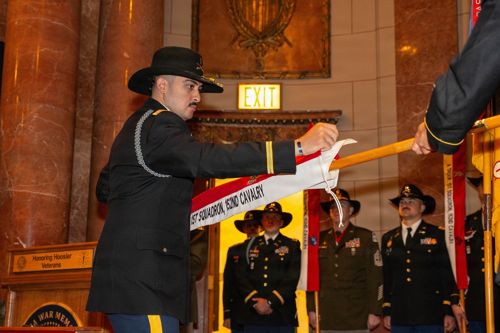 A military ceremony is taking place inside what appears to be the Indiana War Memorial, as indicated by visible signage reading 'Honoring Hoosier Veterans' and 'War Memorial'. A soldier in dress uniform and cavalry Stetson hat is attaching or presenting a streamer reading '1ST SQUADRON, 152ND CAVALRY' to a guidon or unit colors staff. Multiple soldiers in various uniforms stand in formation in the background.