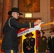 A military ceremony is taking place inside what appears to be the Indiana War Memorial, as indicated by visible signage reading 'Honoring Hoosier Veterans' and 'War Memorial'. A soldier in dress uniform and cavalry Stetson hat is attaching or presenting a streamer reading '1ST SQUADRON, 152ND CAVALRY' to a guidon or unit colors staff. Multiple soldiers in various uniforms stand in formation in the background.