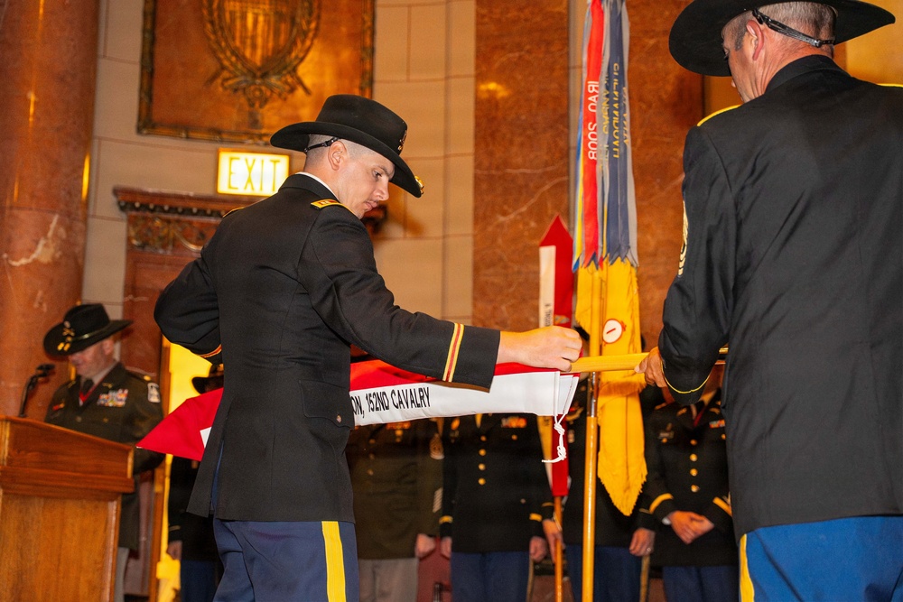 A formal U.S. Army military ceremony is taking place in an ornate indoor venue, likely a state capitol or government building. Two soldiers wearing distinctive black cavalry Stetsons are handling a unit guidon or flag bearing the text '152ND CAVALRY.' Additional soldiers in dress uniforms stand in formation holding flags and streamers in the background.