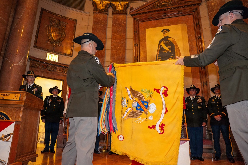 A formal U.S. Army military ceremony is taking place inside an ornate building, likely a state capitol or memorial hall. Two soldiers in dress uniforms are displaying or unfurling a yellow cavalry guidon/regimental flag with streamers, while other uniformed personnel stand at attention in the background. A portrait of a military officer is visible on the wall behind the ceremony.