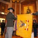 A formal U.S. Army military ceremony is taking place inside an ornate building, likely a state capitol or memorial hall. Two soldiers in dress uniforms are displaying or unfurling a yellow cavalry guidon/regimental flag with streamers, while other uniformed personnel stand at attention in the background. A portrait of a military officer is visible on the wall behind the ceremony.