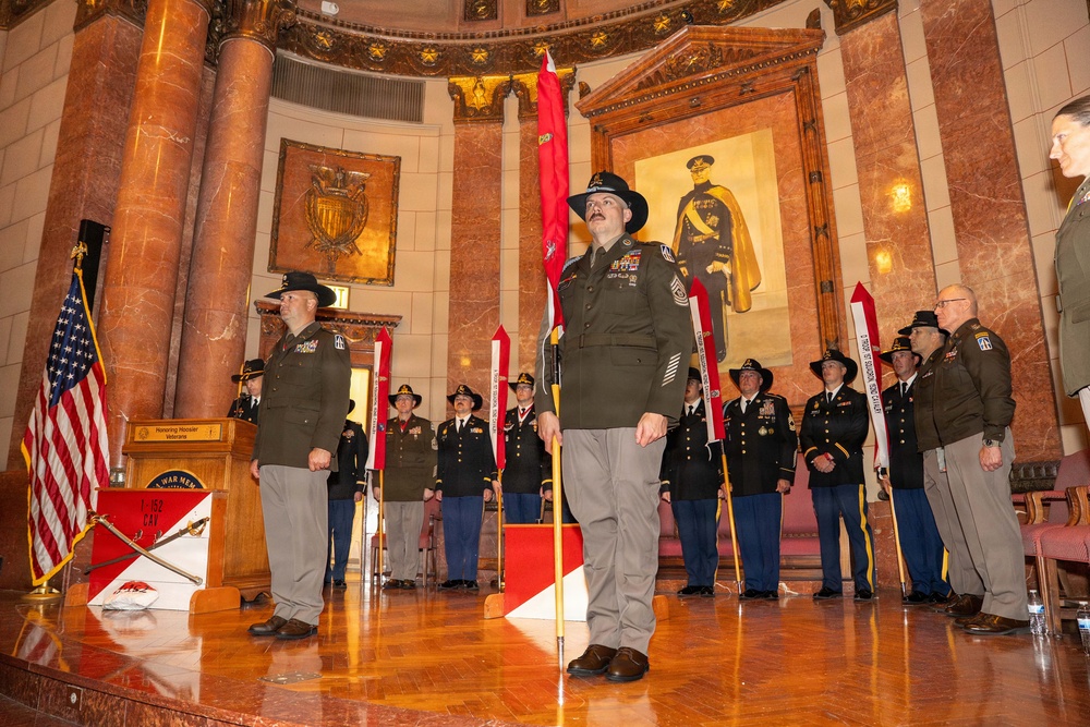 A formal U.S. Army military ceremony taking place inside an ornate rotunda-style hall, likely a war memorial. Soldiers in dress uniforms and campaign hats are standing at attention, with several holding red unit guidon flags. A large portrait of a military officer in historical uniform is visible on the wall behind the formation.