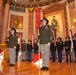 A formal U.S. Army military ceremony taking place inside an ornate rotunda-style hall, likely a war memorial. Soldiers in dress uniforms and campaign hats are standing at attention, with several holding red unit guidon flags. A large portrait of a military officer in historical uniform is visible on the wall behind the formation.