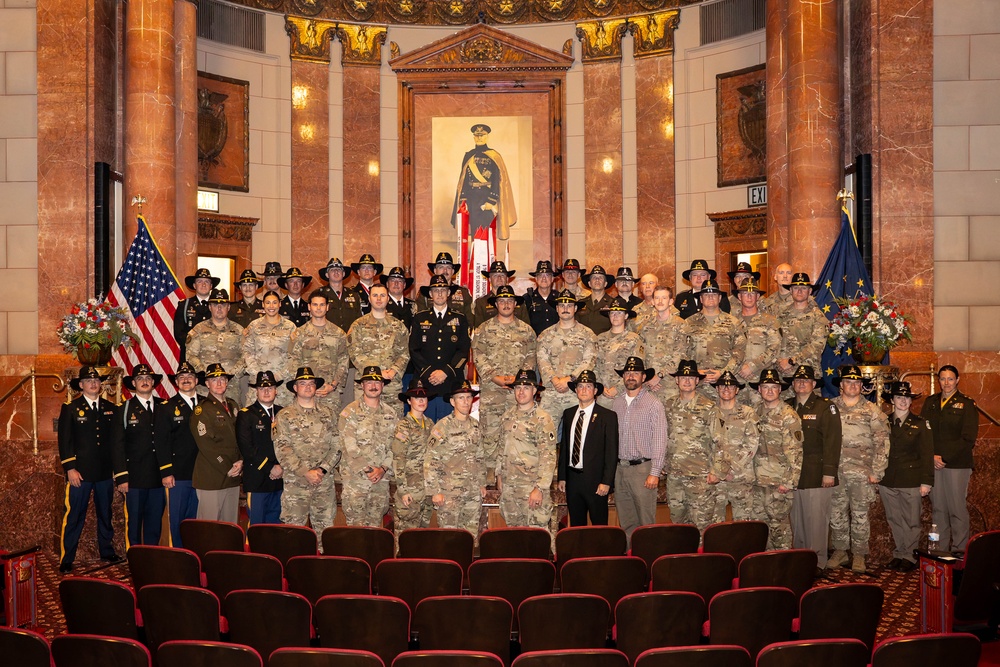 A large group of military personnel and civilians pose for a formal group photograph inside an ornate auditorium or ceremonial hall. The majority of individuals are wearing U.S. Army camouflage uniforms (OCP pattern) with cavalry-style campaign hats, while a smaller number wear dress uniforms. The setting features marble columns, decorative murals, a painted portrait of a military figure, an American flag, and what appears to be a state flag.