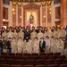 A large group of military personnel and civilians pose for a formal group photograph inside an ornate auditorium or ceremonial hall. The majority of individuals are wearing U.S. Army camouflage uniforms (OCP pattern) with cavalry-style campaign hats, while a smaller number wear dress uniforms. The setting features marble columns, decorative murals, a painted portrait of a military figure, an American flag, and what appears to be a state flag.