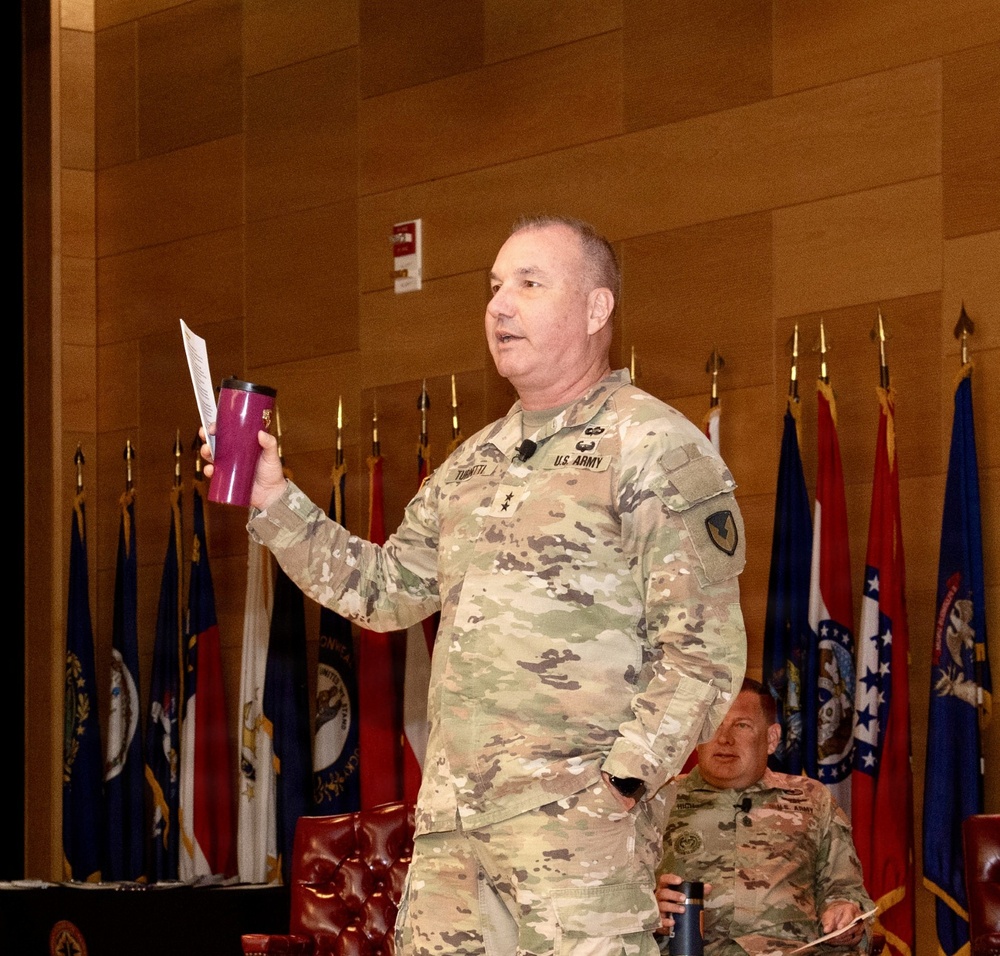 A U.S. Army Major General in OCP (Operational Camouflage Pattern) uniform is speaking at a formal military event or briefing, holding a pink/maroon tumbler and a sheet of paper. Multiple state and military flags are displayed in the background, suggesting a significant command-level gathering. A second uniformed soldier is seated in the background.