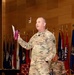 A U.S. Army Major General in OCP (Operational Camouflage Pattern) uniform is speaking at a formal military event or briefing, holding a pink/maroon tumbler and a sheet of paper. Multiple state and military flags are displayed in the background, suggesting a significant command-level gathering. A second uniformed soldier is seated in the background.