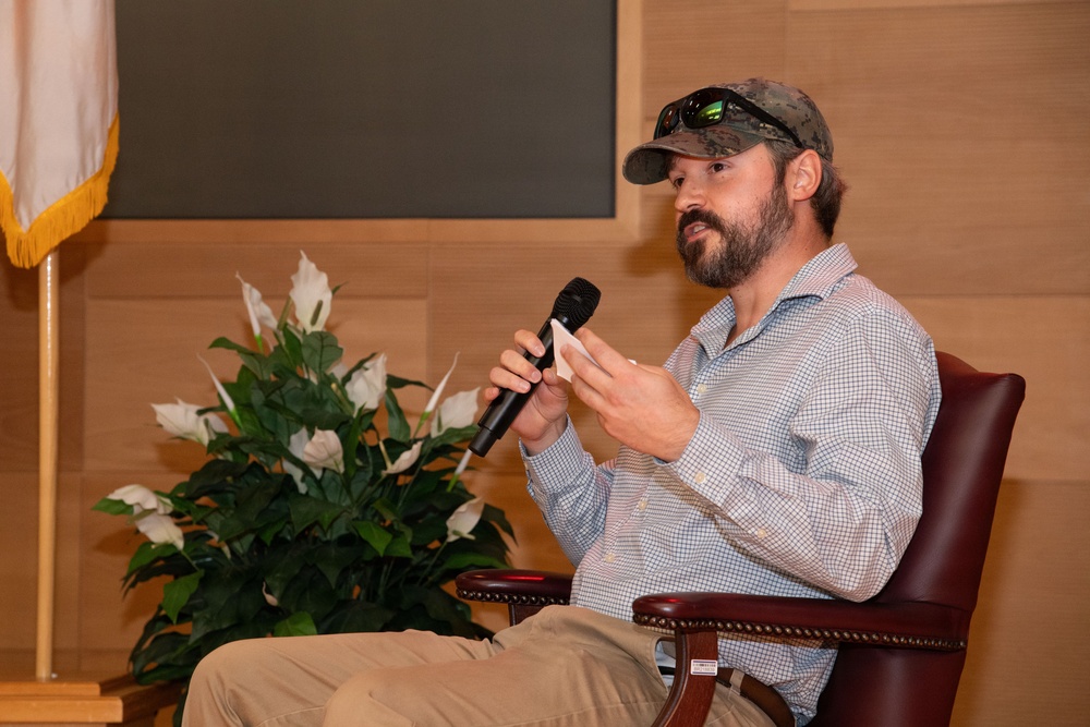 A bearded man wearing a camouflage cap with sunglasses resting on the brim is seated in a leather chair, speaking into a handheld microphone. He is dressed in a light blue checkered button-down shirt and khaki pants, appearing to be a guest speaker or panelist at a formal event. The setting includes a flag with gold fringe, a peace lily plant, and wood-paneled walls suggesting an official or institutional venue.