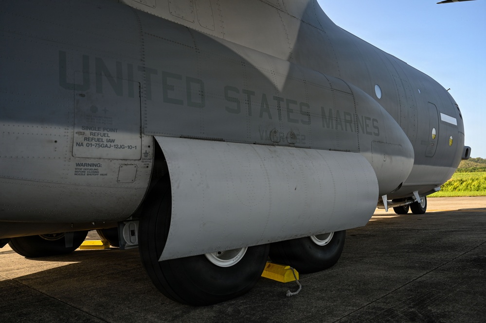 The image shows the fuselage and landing gear section of a United States Marines military transport aircraft parked on a tarmac. The aircraft displays stenciled text reading 'UNITED STATES MARINES' along with maintenance markings including single-point refuel instructions. Yellow wheel chocks are visible securing the main landing gear bogies.