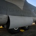 The image shows the fuselage and landing gear section of a United States Marines military transport aircraft parked on a tarmac. The aircraft displays stenciled text reading 'UNITED STATES MARINES' along with maintenance markings including single-point refuel instructions. Yellow wheel chocks are visible securing the main landing gear bogies.