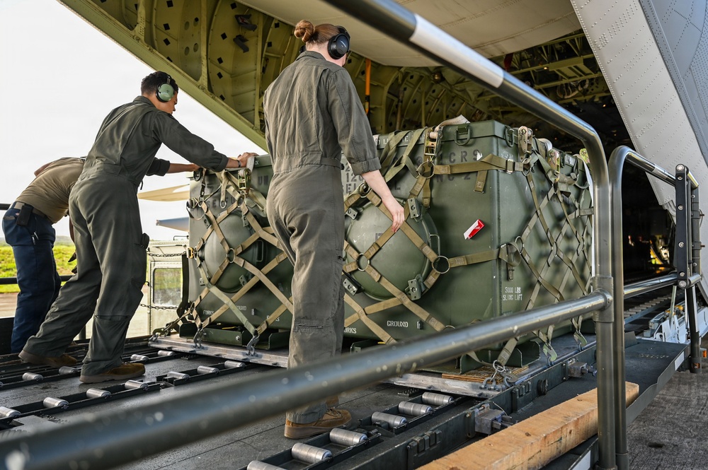 Military personnel in olive drab flight suits are loading or unloading cargo onto a military transport aircraft via a roller-equipped cargo ramp. The cargo consists of military equipment secured in netting and straps on a standard military pallet. At least three personnel are visible, wearing hearing protection, actively maneuvering the pallet.