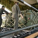 Military personnel in olive drab flight suits are loading or unloading cargo onto a military transport aircraft via a roller-equipped cargo ramp. The cargo consists of military equipment secured in netting and straps on a standard military pallet. At least three personnel are visible, wearing hearing protection, actively maneuvering the pallet.