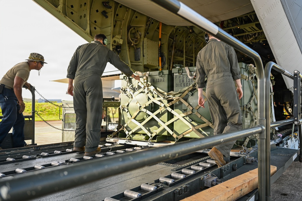 Military personnel are loading cargo onto a large military transport aircraft via its rear ramp. Three individuals in military attire are guiding a strapped pallet of equipment — marked with 'UN 1202 Diesel' — onto roller tracks inside the aircraft. The operation appears to be taking place on a tarmac or airfield.