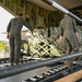Military personnel are loading cargo onto a large military transport aircraft via its rear ramp. Three individuals in military attire are guiding a strapped pallet of equipment — marked with 'UN 1202 Diesel' — onto roller tracks inside the aircraft. The operation appears to be taking place on a tarmac or airfield.