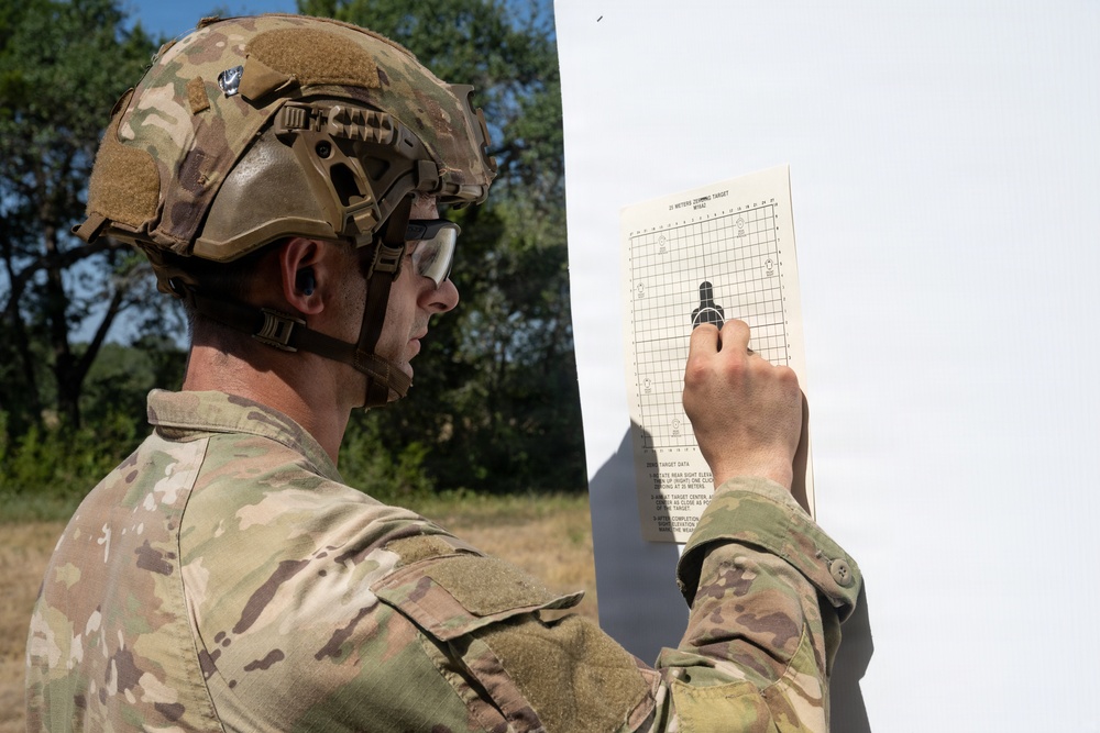 A soldier in full camouflage uniform and tactical helmet is examining or marking a military zeroing target sheet mounted on a white surface. The target appears to be a standard 25-meter zeroing target used for rifle sight alignment. The soldier is holding a pen or marker and appears to be recording shot group data on the target.