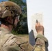 A soldier in full camouflage uniform and tactical helmet is examining or marking a military zeroing target sheet mounted on a white surface. The target appears to be a standard 25-meter zeroing target used for rifle sight alignment. The soldier is holding a pen or marker and appears to be recording shot group data on the target.