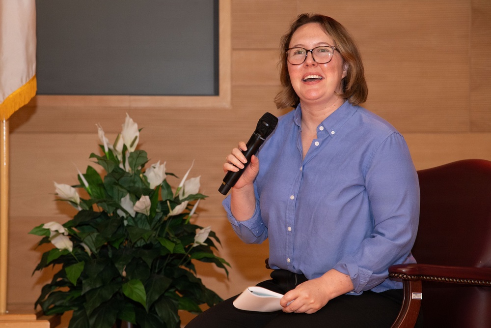 A woman with shoulder-length blonde hair and black-rimmed glasses is seated in a burgundy leather chair, holding a black handheld microphone and smiling broadly. She is wearing a blue button-down shirt and holding papers in her other hand, suggesting she is speaking at a formal or semi-formal event. The setting appears to be an auditorium or meeting hall with wood-paneled walls and a decorative peace lily plant nearby.