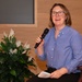 A woman with shoulder-length blonde hair and black-rimmed glasses is seated in a burgundy leather chair, holding a black handheld microphone and smiling broadly. She is wearing a blue button-down shirt and holding papers in her other hand, suggesting she is speaking at a formal or semi-formal event. The setting appears to be an auditorium or meeting hall with wood-paneled walls and a decorative peace lily plant nearby.
