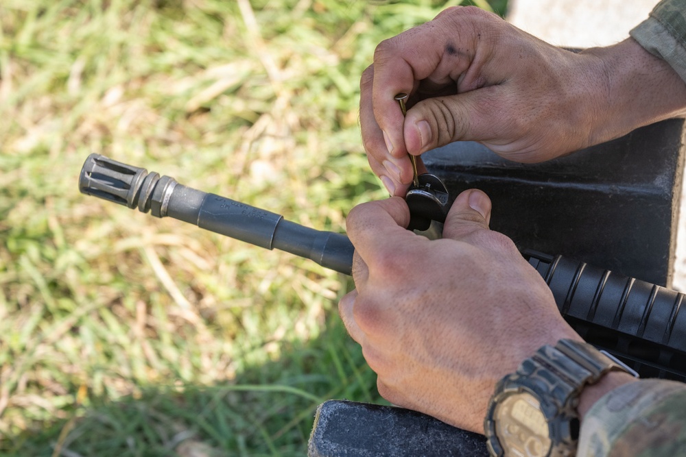 A person is handling what appears to be a rifle, specifically working near the bolt carrier or upper receiver area while holding a small pin or tool. The firearm has a visible flash hider/muzzle device on the barrel end and a handguard with rail sections. The activity appears to be field maintenance or assembly/disassembly of the weapon.