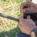 A person is handling what appears to be a rifle, specifically working near the bolt carrier or upper receiver area while holding a small pin or tool. The firearm has a visible flash hider/muzzle device on the barrel end and a handguard with rail sections. The activity appears to be field maintenance or assembly/disassembly of the weapon.