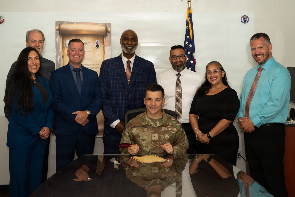 A U.S. Air Force officer in military camouflage uniform, identified by name tape as 'Wilson' and wearing a general's star, is seated at a desk signing or presenting an official document. Seven civilians in business attire stand behind him in what appears to be an official ceremony or signing event. An American flag and organizational posters are visible in the background.