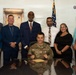 A U.S. Air Force officer in military camouflage uniform, identified by name tape as 'Wilson' and wearing a general's star, is seated at a desk signing or presenting an official document. Seven civilians in business attire stand behind him in what appears to be an official ceremony or signing event. An American flag and organizational posters are visible in the background.