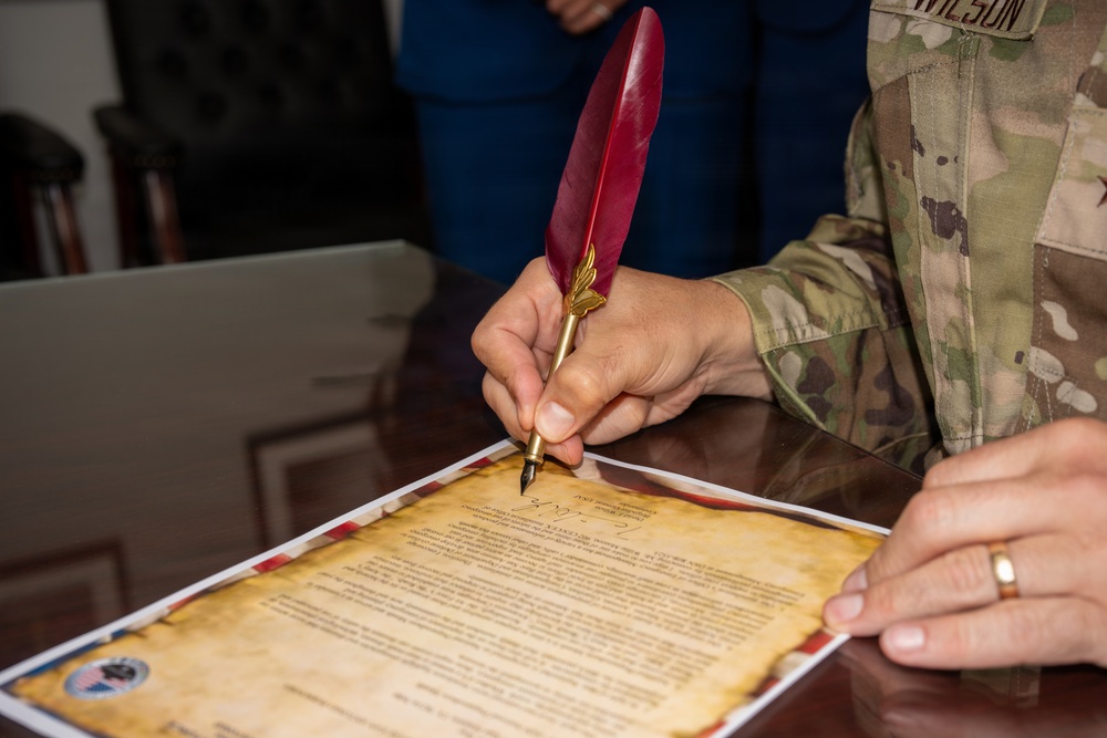 A person in military camouflage uniform (name tape reading 'WILSON' visible) is signing an official document using a decorative red quill pen with a gold nib and gold accent piece. The document appears to be a formal certificate or proclamation printed on aged/parchment-style paper with decorative borders. A second person in civilian clothing (blue) is present in the background.