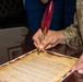 A person in military camouflage uniform (name tape reading 'WILSON' visible) is signing an official document using a decorative red quill pen with a gold nib and gold accent piece. The document appears to be a formal certificate or proclamation printed on aged/parchment-style paper with decorative borders. A second person in civilian clothing (blue) is present in the background.