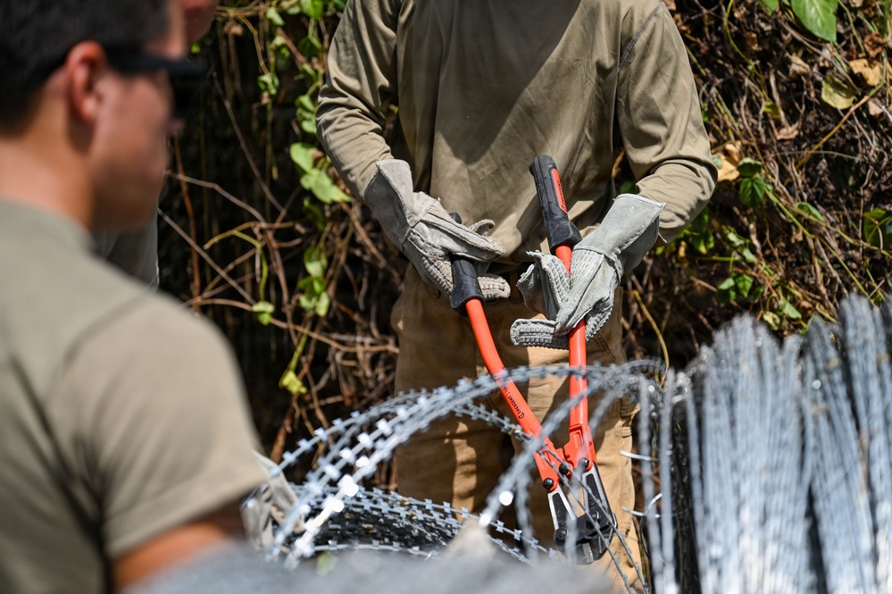 Two individuals are working with concertina/razor wire in an outdoor setting with vegetation in the background. The central figure is wearing work gloves and holding red-handled bolt cutters, apparently cutting or handling the coiled razor wire. A second person is partially visible on the left side of the frame.