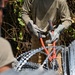 Two individuals are working with concertina/razor wire in an outdoor setting with vegetation in the background. The central figure is wearing work gloves and holding red-handled bolt cutters, apparently cutting or handling the coiled razor wire. A second person is partially visible on the left side of the frame.