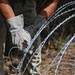 A person wearing protective work gloves is handling concertina (razor) wire, appearing to install or deploy it in an outdoor wooded area. The individual is dressed in olive/military-style clothing and is using a small tool, possibly zip ties or wire fasteners, to secure the coiled razor wire. The setting appears to be a perimeter security or barrier installation task.
