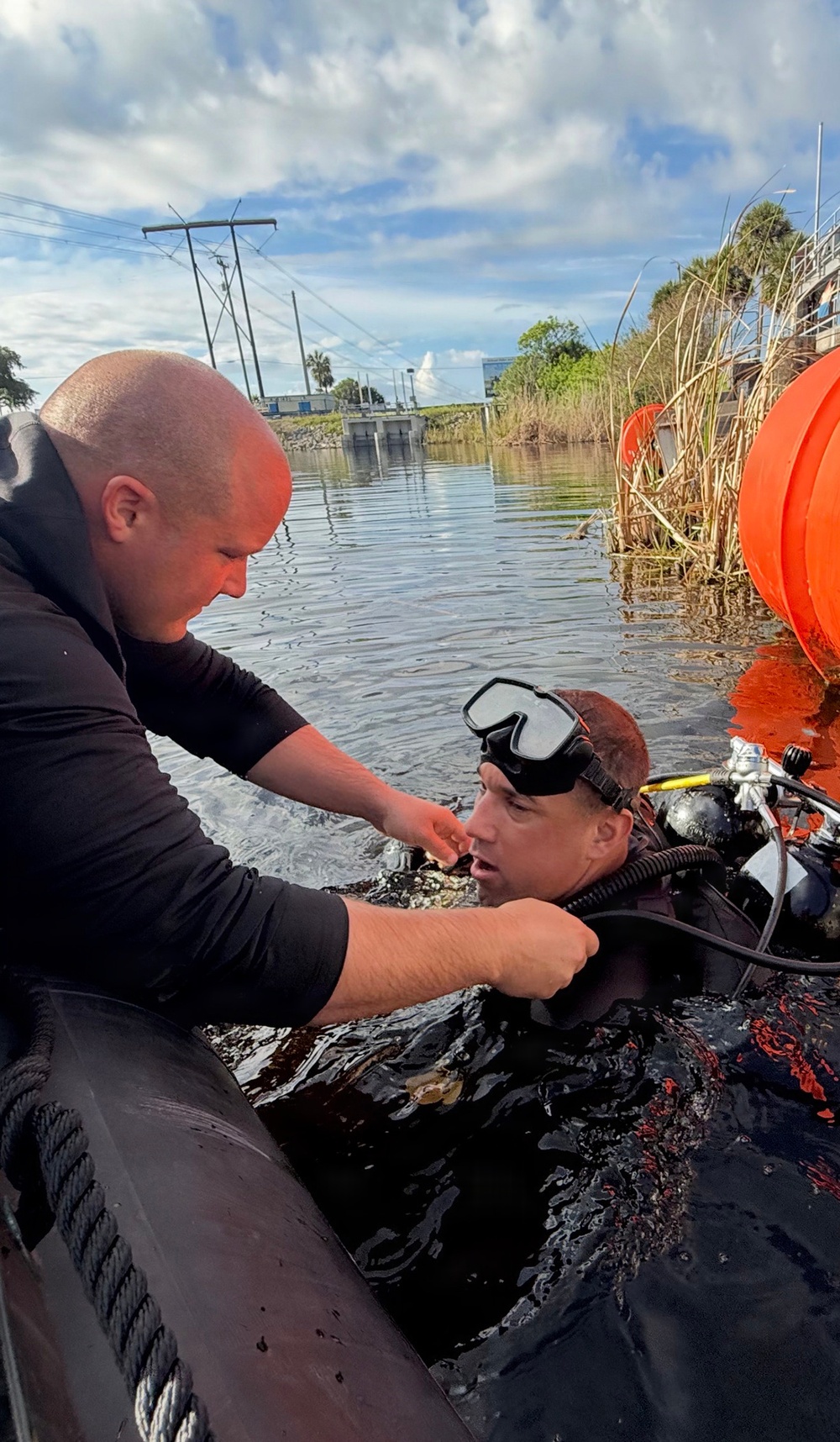 Two individuals are engaged in a diving operation near a waterway. One person in a black long-sleeve shirt is assisting a fully equipped scuba diver who is partially submerged in dark water. The setting appears to be a canal or retention pond with utility infrastructure visible in the background.