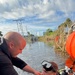 Two individuals are engaged in a diving operation near a waterway. One person in a black long-sleeve shirt is assisting a fully equipped scuba diver who is partially submerged in dark water. The setting appears to be a canal or retention pond with utility infrastructure visible in the background.